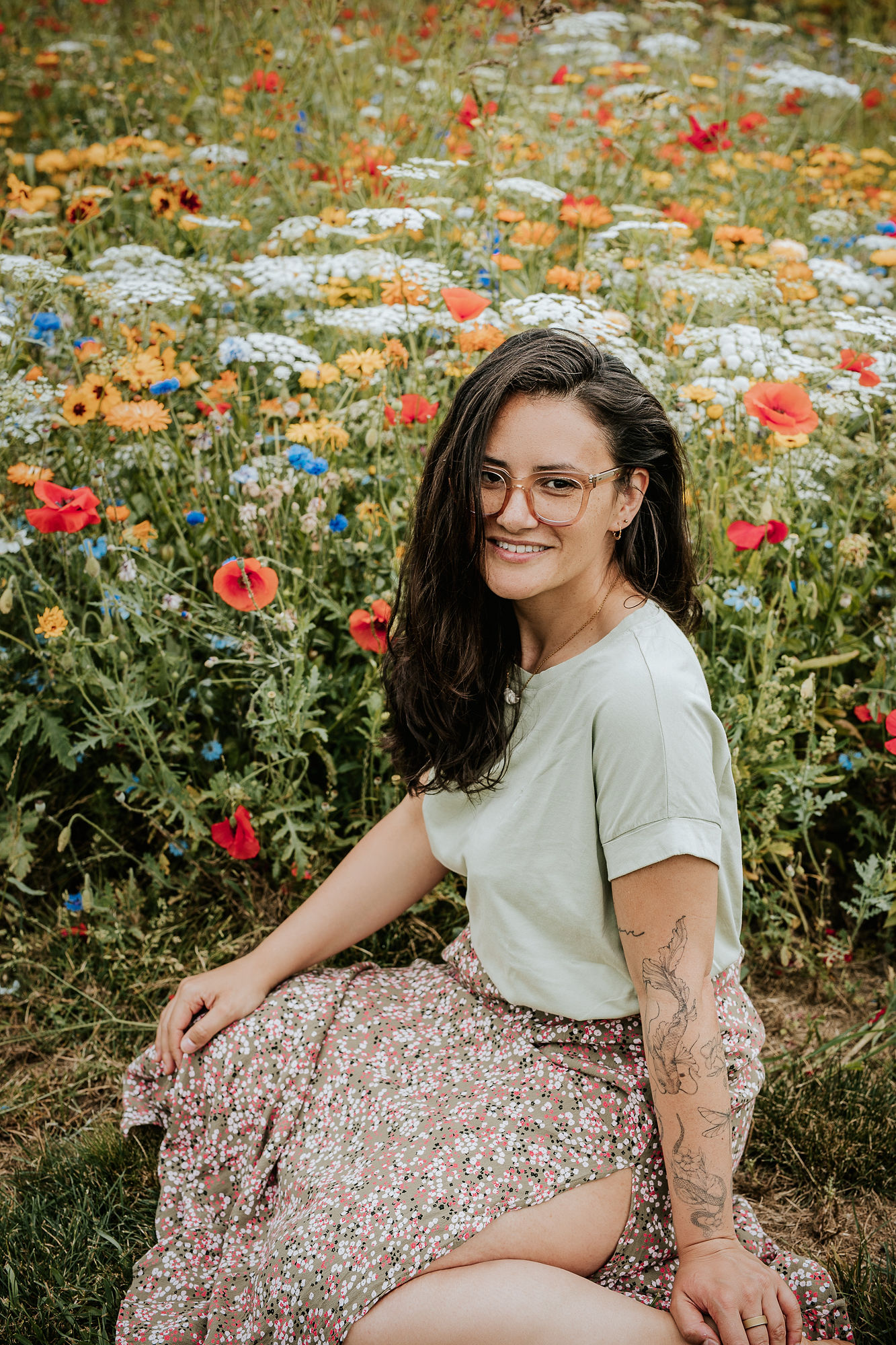 picture of steffi von brunner, she sits in easy pose on the ground and looks straight to the camera. She wears a trousers and a wrap top in a black and white flower print and wears pink transluscent glasses
