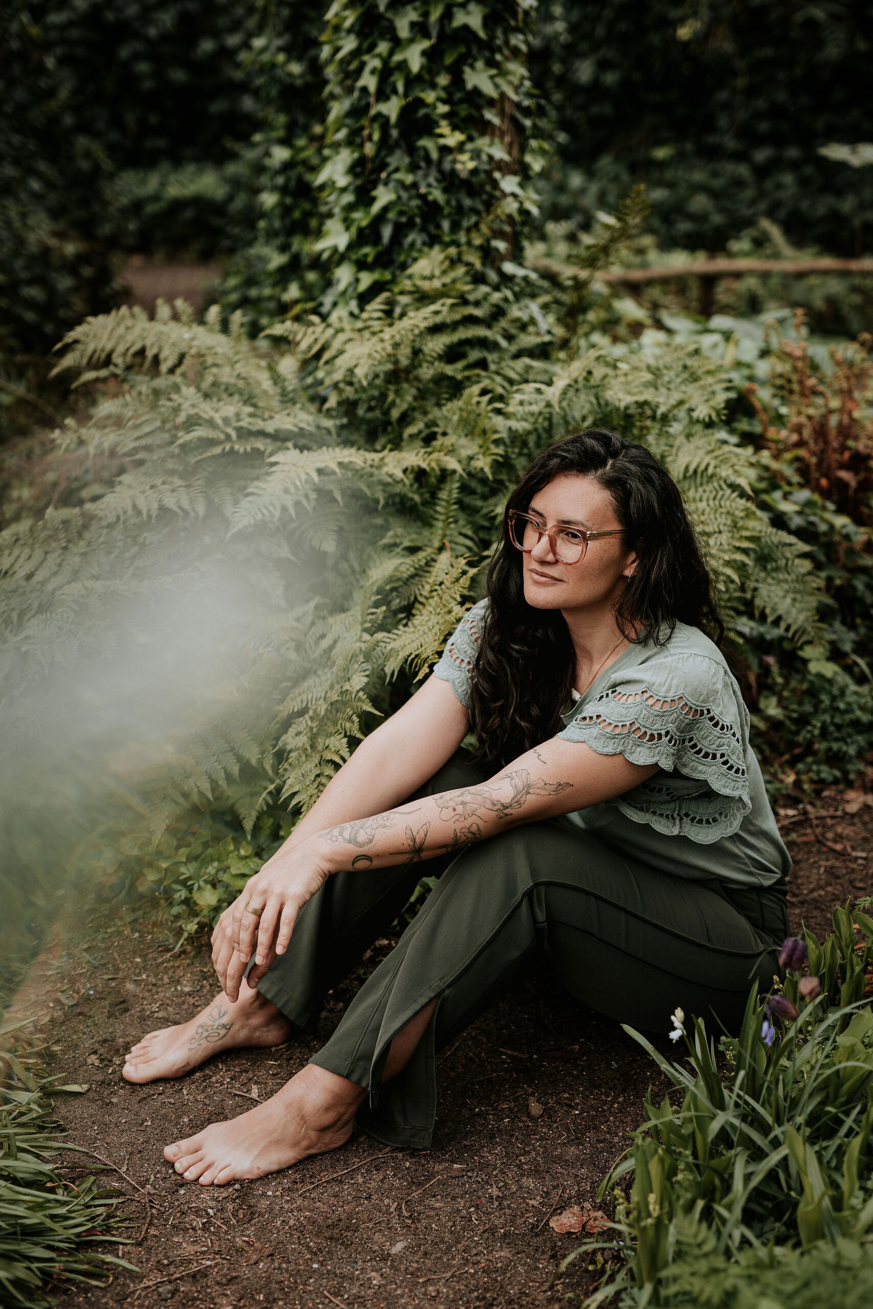 picture of steffi von brunner, she sits in easy pose on the ground and looks straight to the camera. She wears a trousers and a wrap top in a black and white flower print and wears pink transluscent glasses