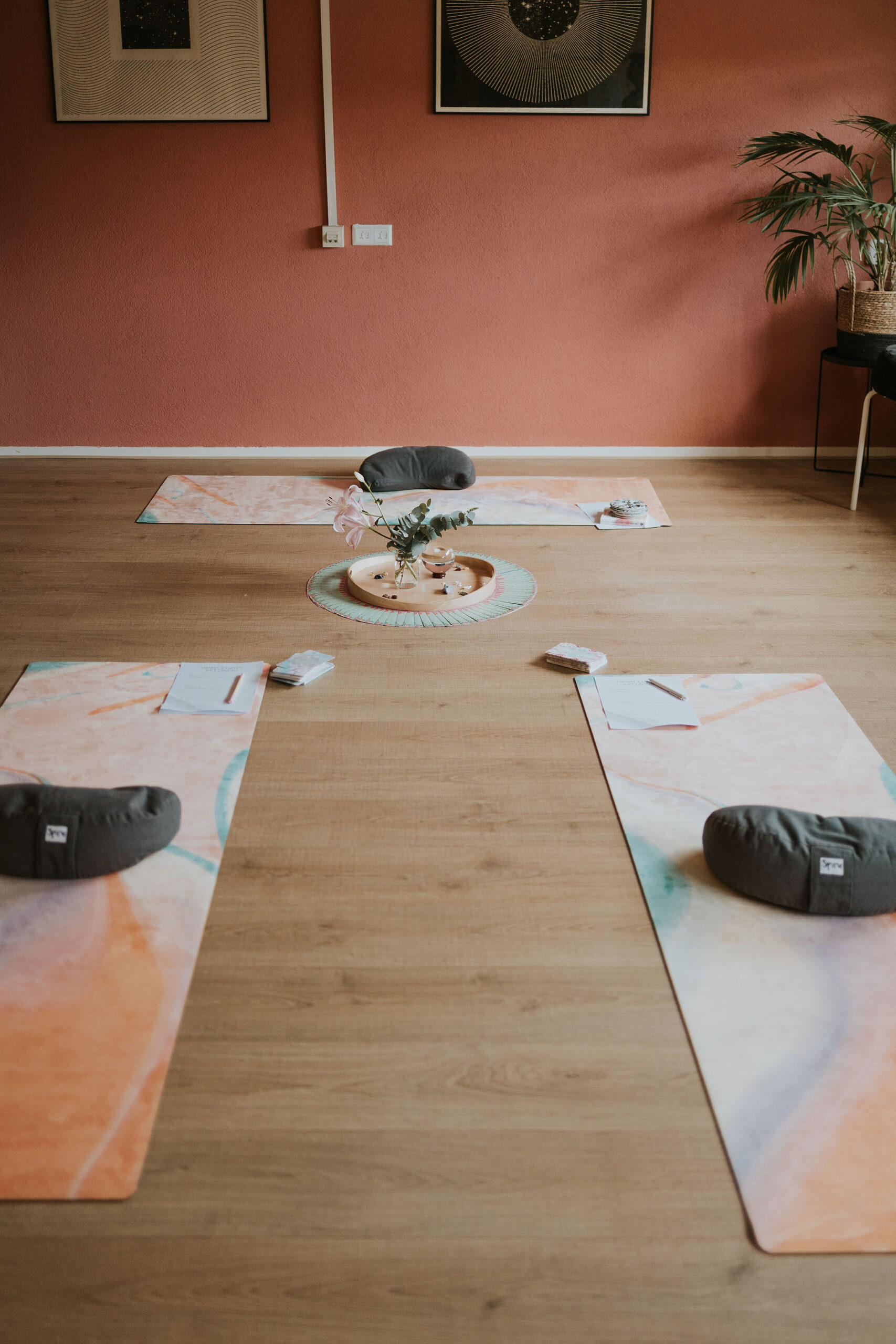 20260704_STEFFI_fotograafLANDAPENDERS_009 Yogamatten met meditatiekussens liggen in een cirkel klaar voor een groepssessie genaamd Soft Ground, met werkbladen en pennen erop en een altaartje met bloemen en kristallen in het midden. De ruimte heeft een terracotta muur en zonachtig kunstwerk op de achtergrond.