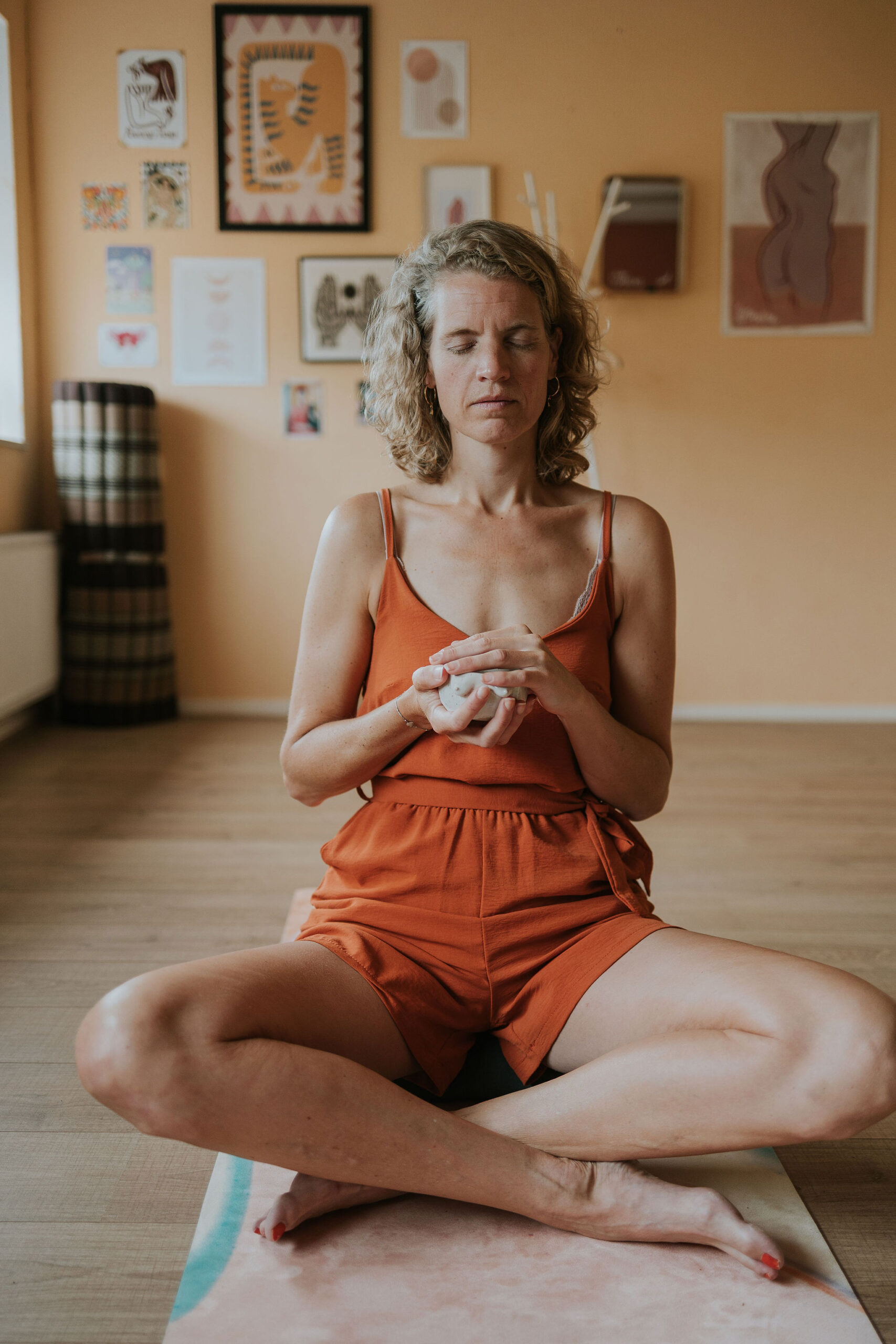 20260704_STEFFI_fotograafLANDAPENDERS_017 Een vrouw zit met gesloten ogen op een yogamat en houdt een kopje cacao met beide handen tegen haar hart, met een muur vol illustraties op de achtergrond tijdens een groep sessie genaamd soft ground van Steffi von Brunner