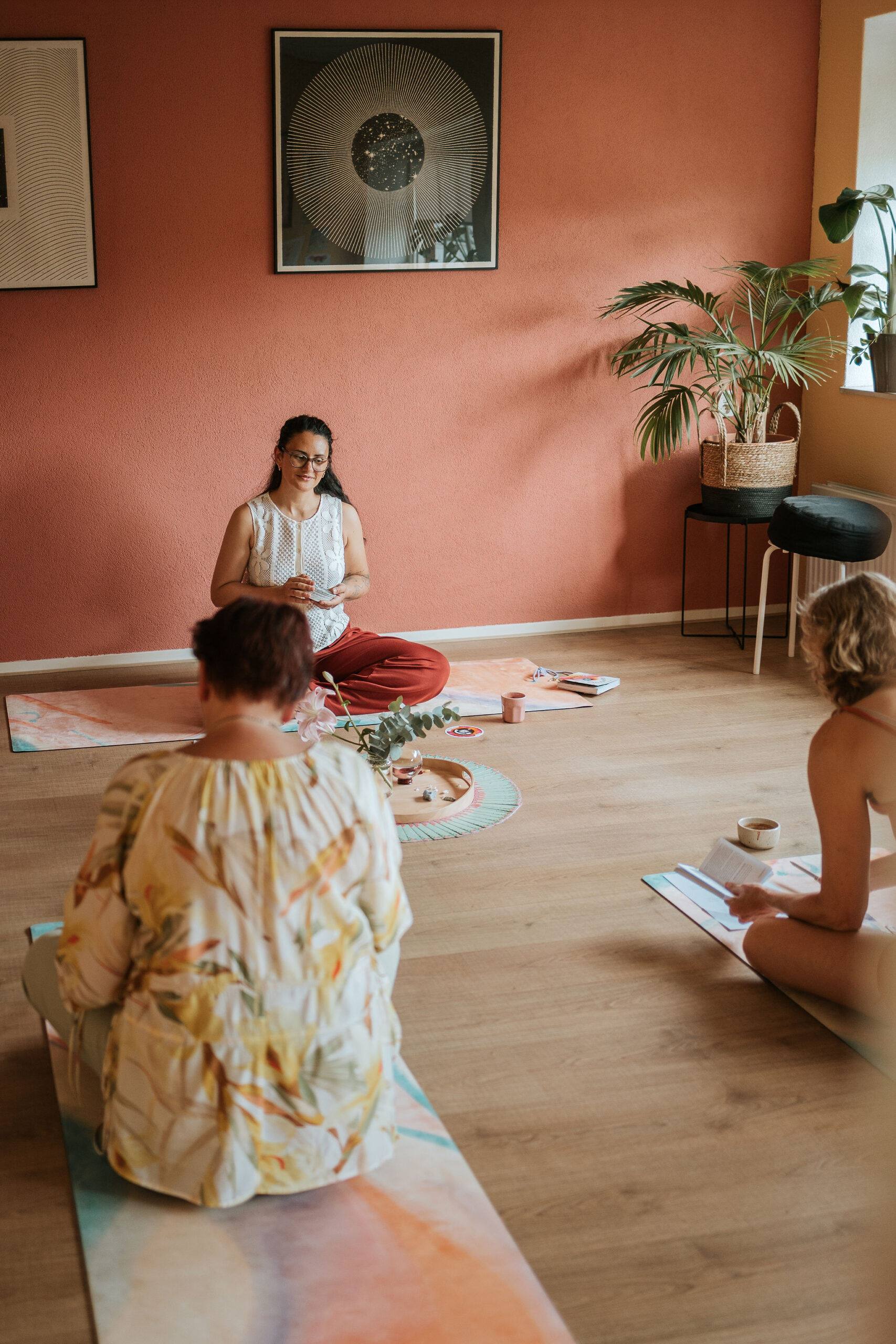 20260704_STEFFI_fotograafLANDAPENDERS_023 Steffi zit met gekruiste benen op een yogamat en houdt kaarten vast, terwijl twee vrouwen om haar heen zitten tijdens een groepssessie genaamd Soft Ground. Op de grond ligt een altaartje met bloemen tussen de matten in, met een terracotta muur en zonachtig kunstwerk op de achtergrond.