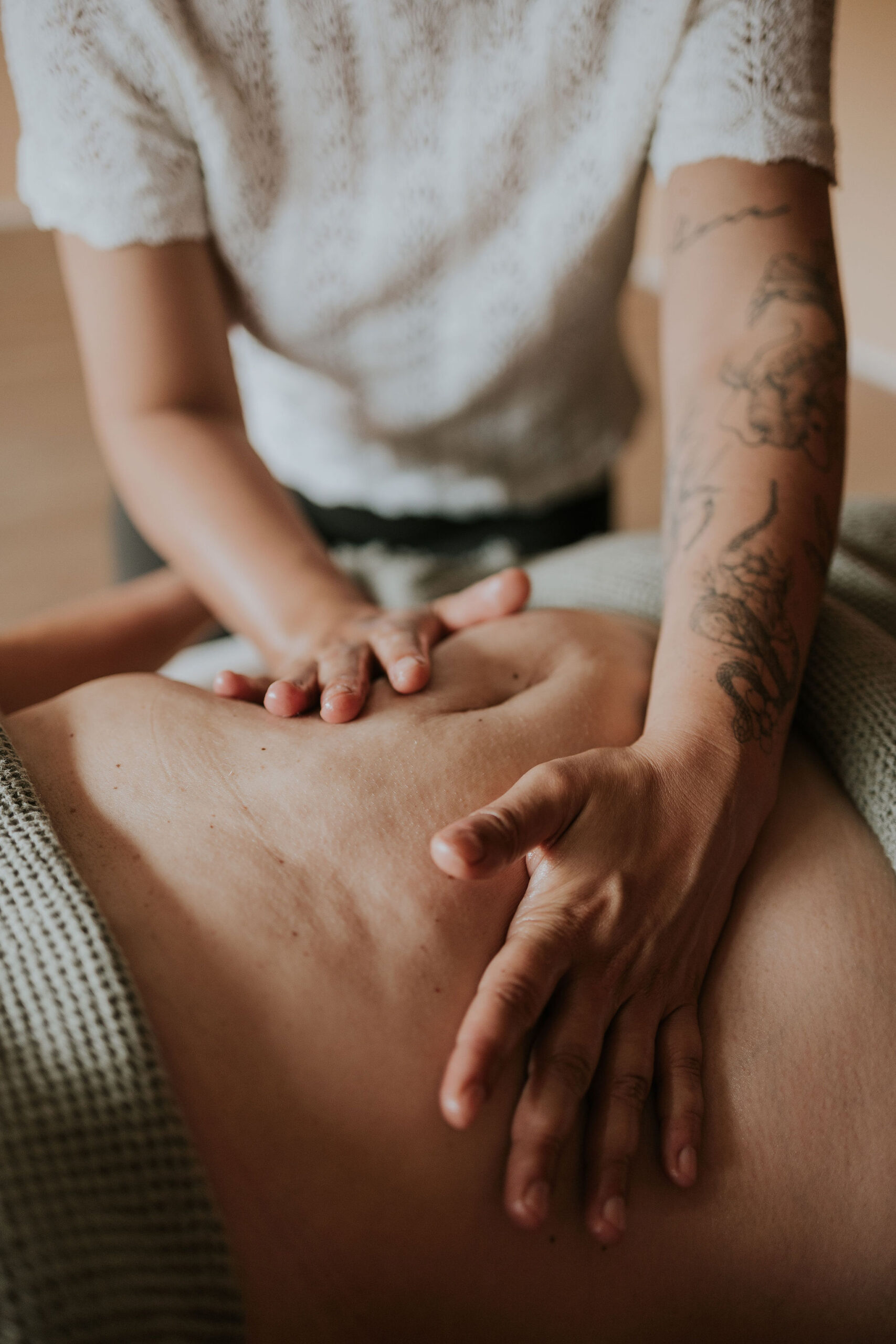 Close up van twee handen die rusten op een blote buik tijdens een Chi Nei Tsang behandeling, met een getatoeëerde onderarm en een groene deken op de achtergrond.
